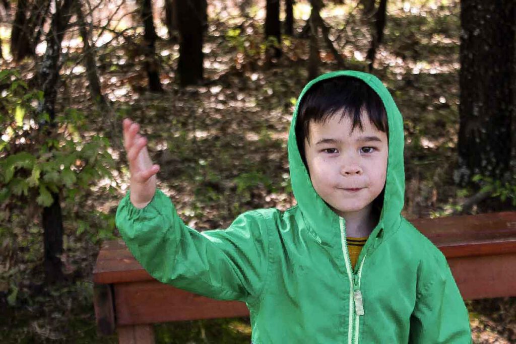 A young boy in a green hooded jacket raises his hand outdoors on a wooded trail.