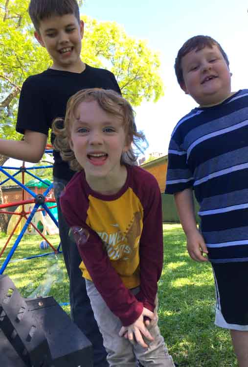 Three smiling boys playing outdoors with a bubble machine beside a climbing dome at Bright Mosaic.