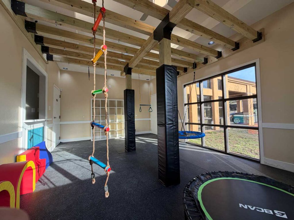 A custom-built indoor gym at Bright Mosaic in Sherman, TX, featuring a climbing rope ladder, trampoline, swing, and padded pillars for safe play.