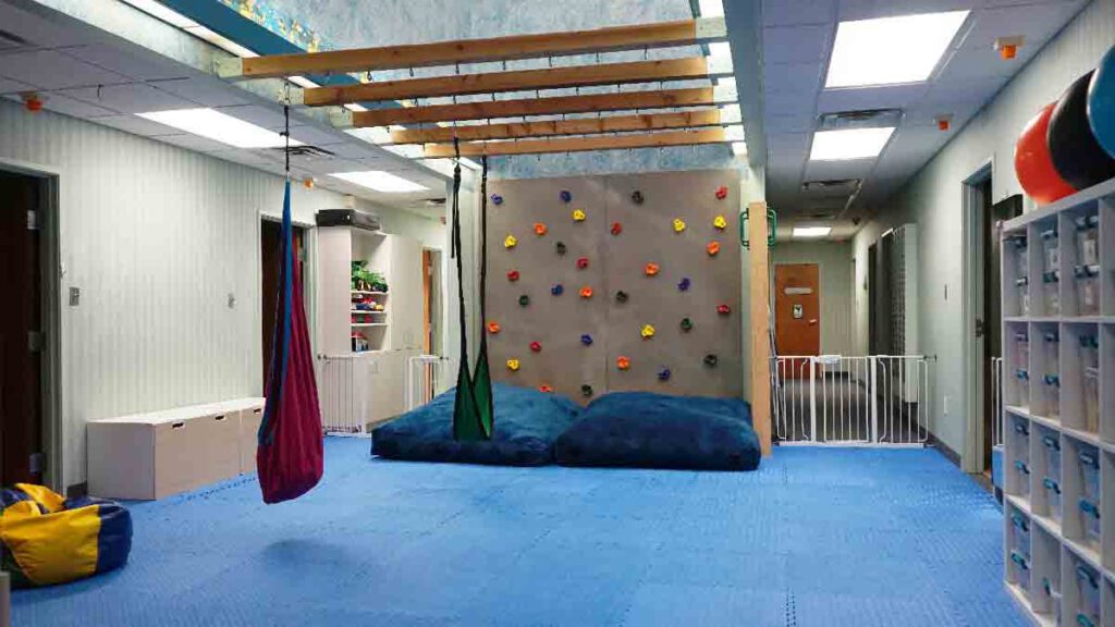 Large indoor therapy gym at Bright Mosaic on Bonnie Brae St. featuring a colorful rock climbing wall, overhead wooden monkey bars, sensory swings, and blue foam safety flooring for pediatric ABA and OT.