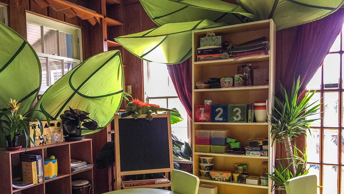 Therapeutic art and play room at Bright Mosaic in Denton, featuring a jungle theme with large leaf canopies, wooden shelves with educational toys, and a chalkboard for creative ABA therapy.