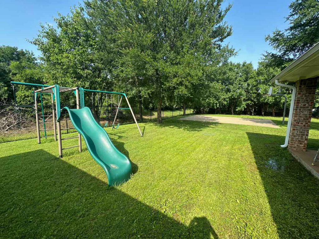 Large private outdoor play area at Bright Mosaic Southlake featuring multiple green slides, a swing set, a monkey bar structure, and a concrete basketball court surrounded by a secure fence and mature trees.