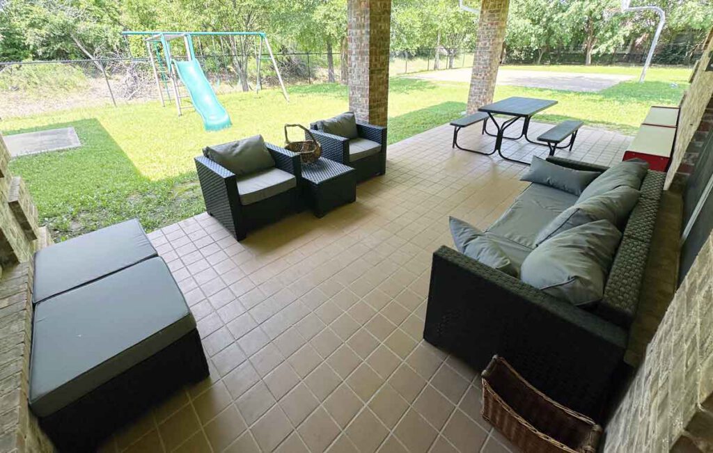 Shaded outdoor patio at Bright Mosaic Southlake featuring modern wicker lounge seating, a picnic table, and a view of the secure grassy play area with a slide and swing set.