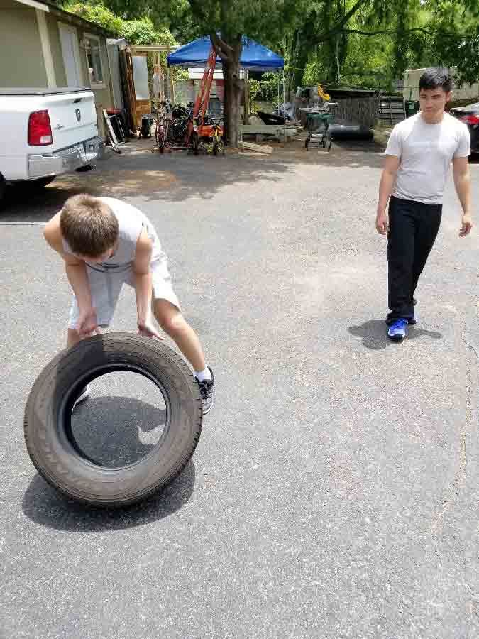 A young boy flips a large tire outdoors while his occupational therapist watches to support his gross motor development.