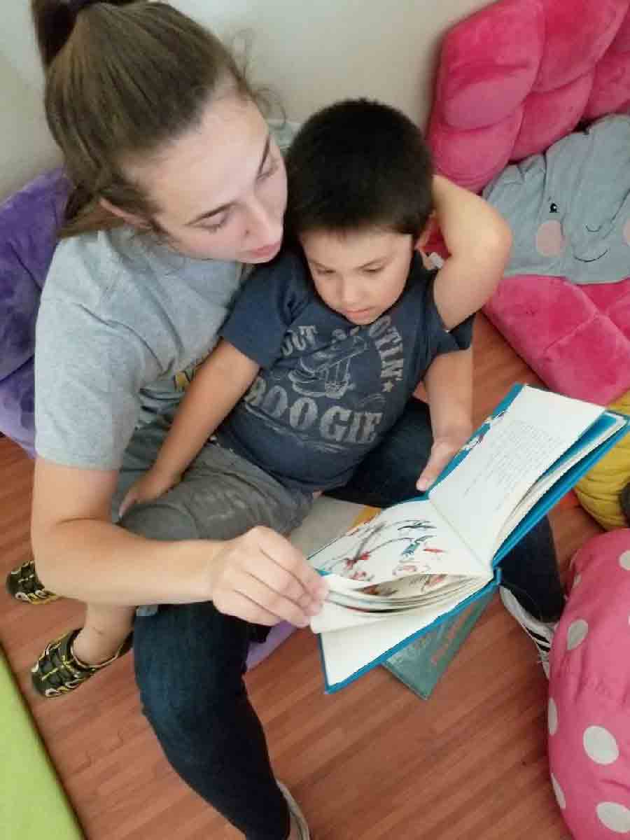 A pediatric therapist sits on the floor reading a picture book with a young boy during a personalized autism therapy session.