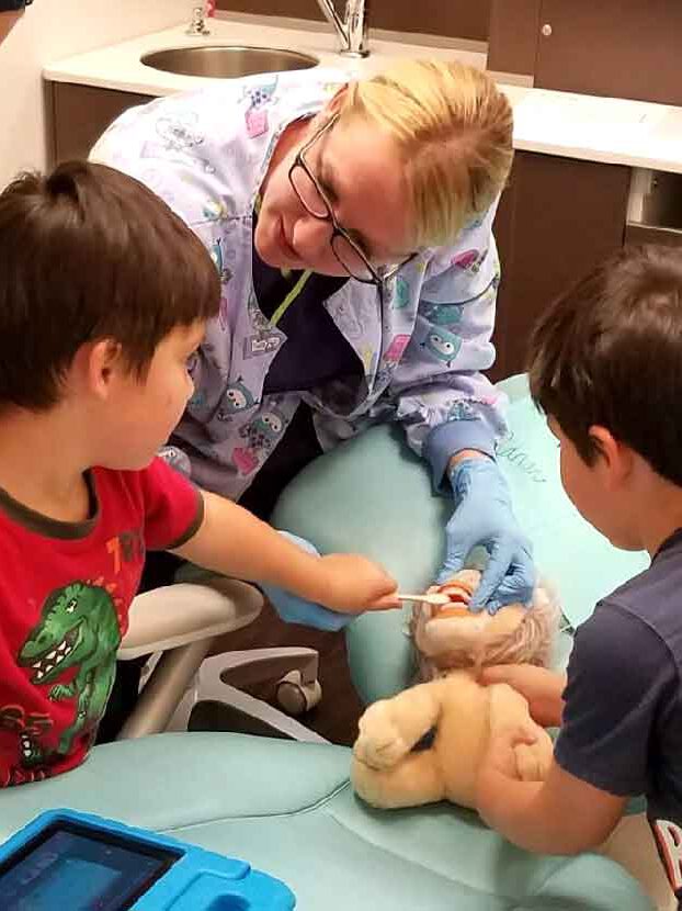 Two young boys practice brushing a stuffed animal's teeth during a dentist office visit as part of an occupational therapy life skills program.