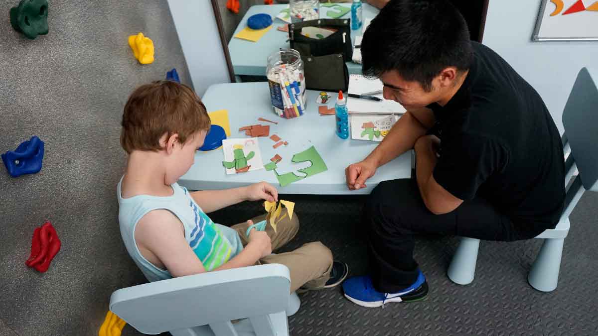 A young boy sitting at a table and practicing fine motor skills by cutting paper shapes with scissors while guided by a pediatric occupational therapist.