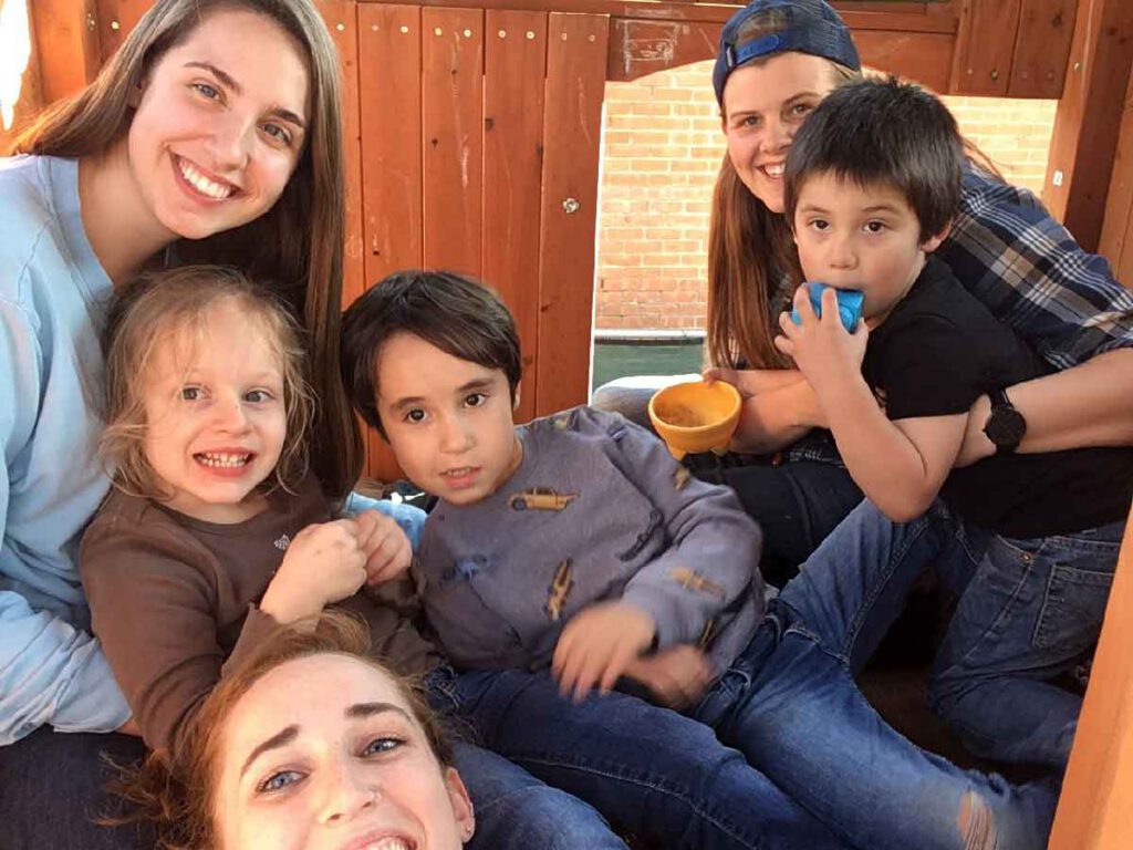 Three smiling Bright Mosaic team members sit with three young children inside a wooden playground fort during a play-based session.